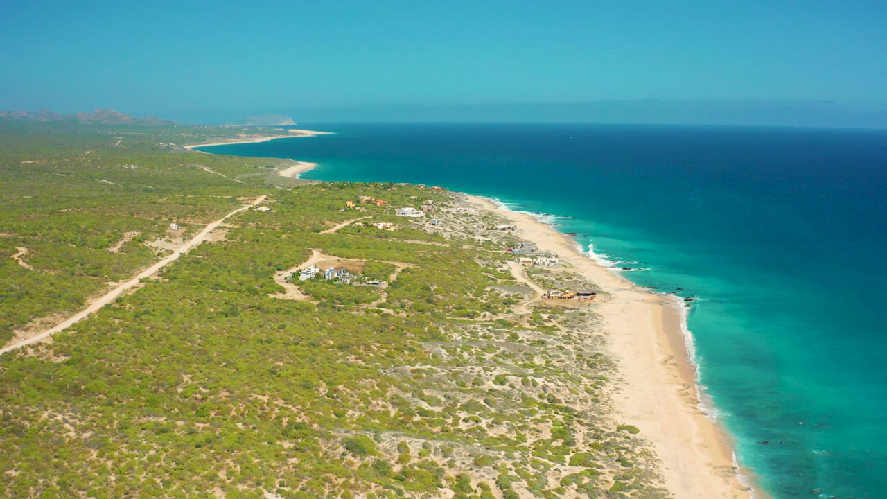 Opening aerial frame over El Cardoncito, Baja California Sur, showing a diagonal sand beach, turquoise shoreline, green scrubland, sandy tracks, distant hills, and no visible people.