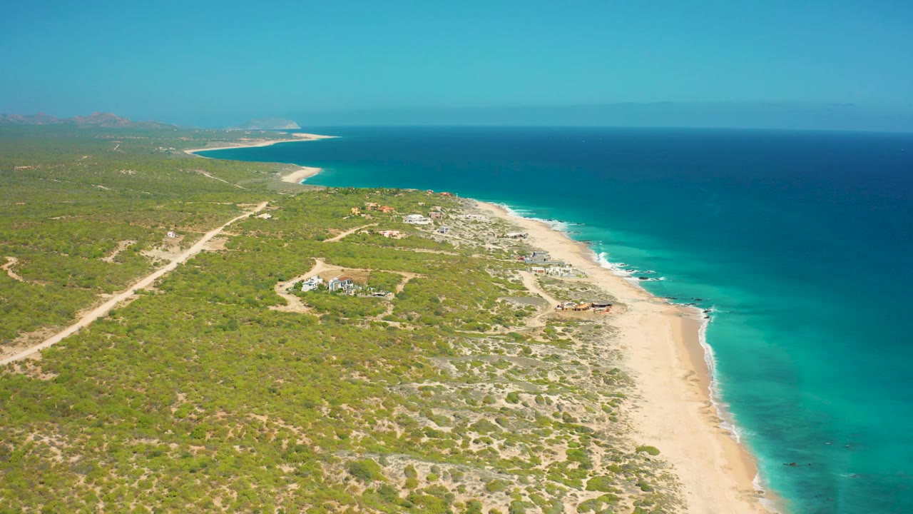 High aerial frame over El Cardoncito showing the long beach on the right edge, layered turquoise and dark blue water, low green vegetation inland, sandy tracks, distant mountains, and no visible people.