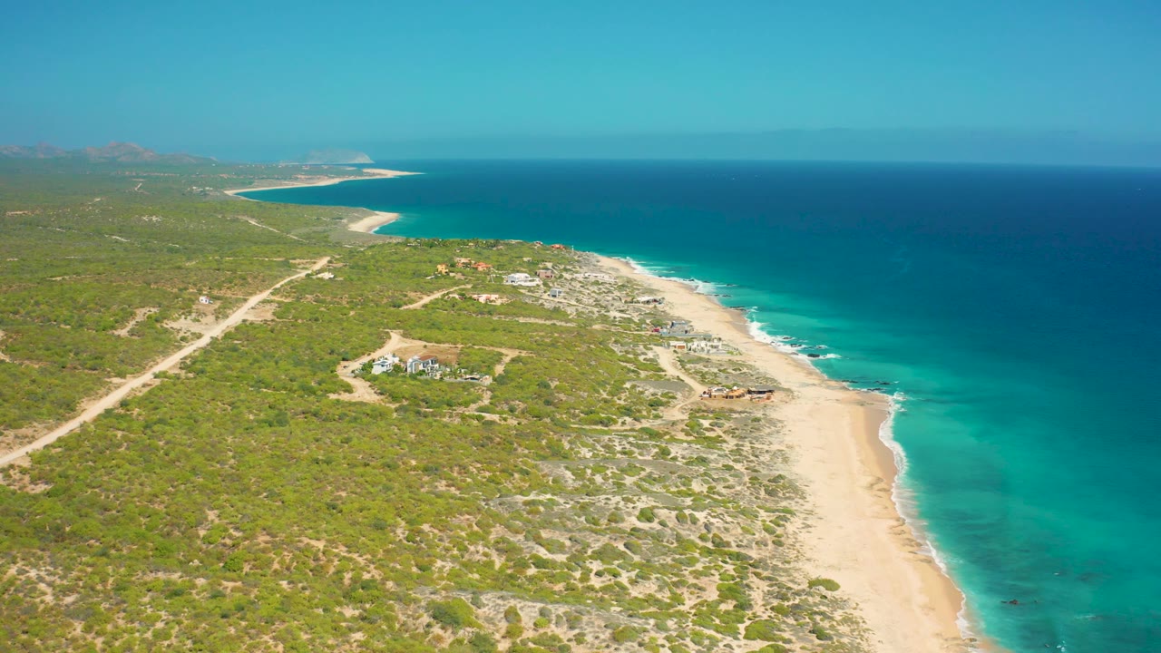 Aerial coastline with a pale beach, green brush, sandy road, and open blue sea.