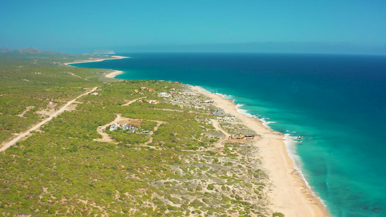 Aerial view over El Cardoncito, Baja California Sur, with turquoise water, pale sand, scrub-covered land, dirt tracks, scattered buildings, distant headlands, and no visible people.