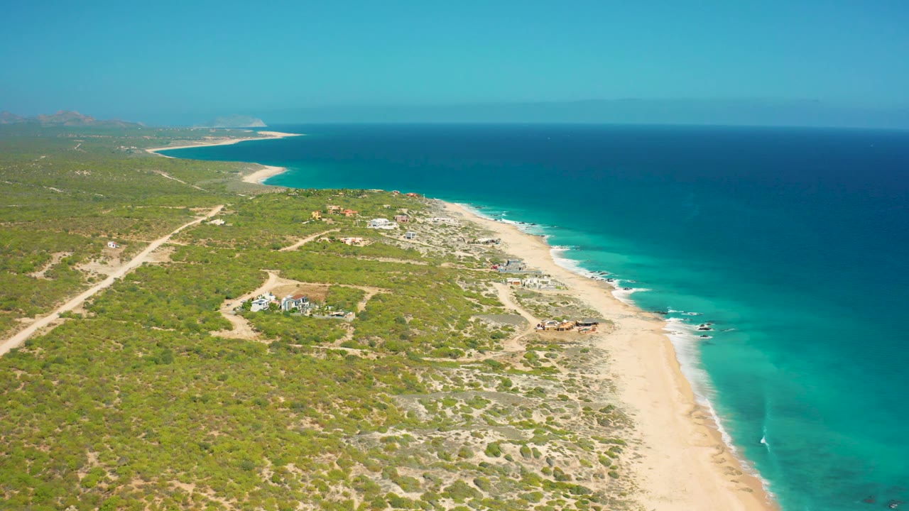 Aerial frame over El Cardoncito showing turquoise shallows against the sand, darker ocean beyond, inland tracks through low vegetation, a few buildings near the coast, and no visible people.