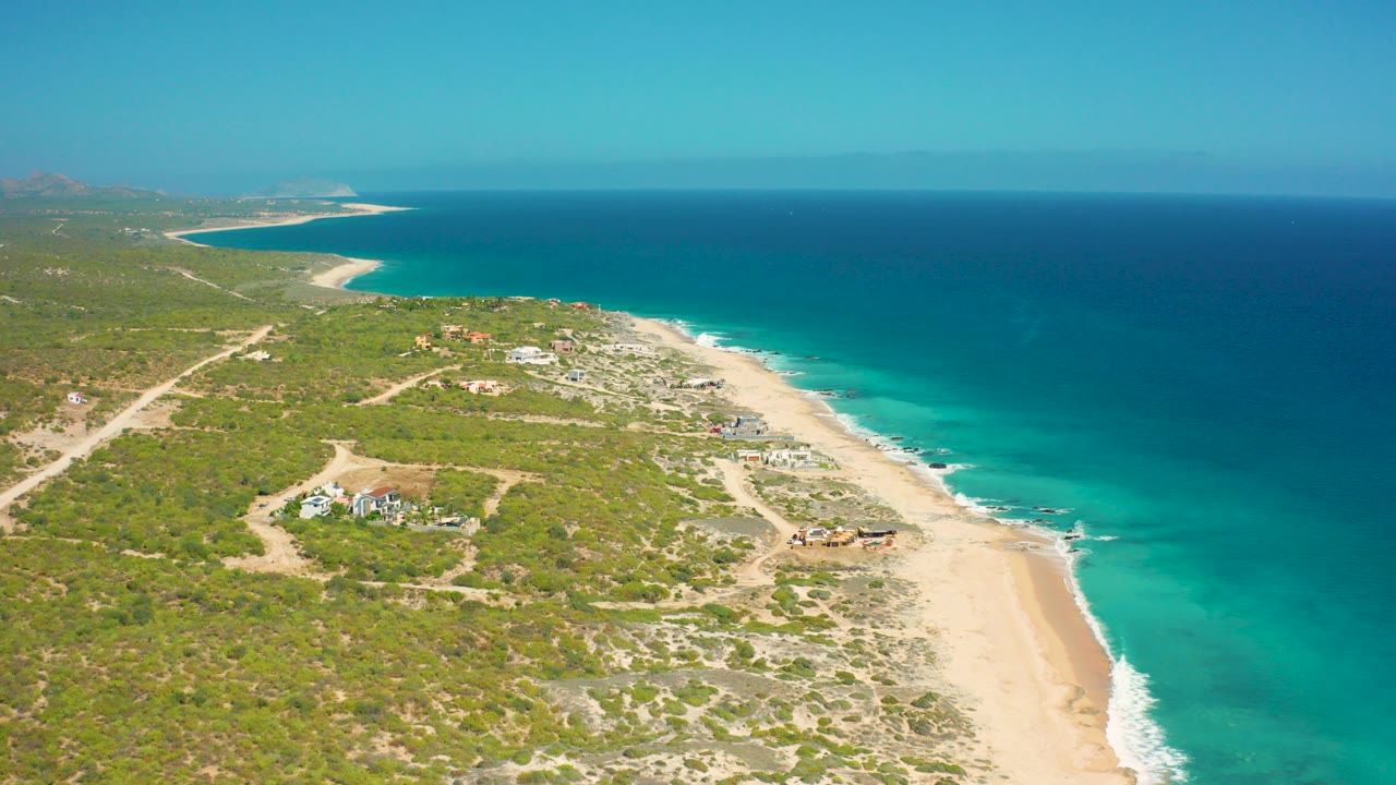 A high aerial view over El Cardoncito, Baja California Sur, Mexico shows scrubland and dirt tracks on the left, a pale beach curving along turquoise water, darker sea beyond, and no visible people.