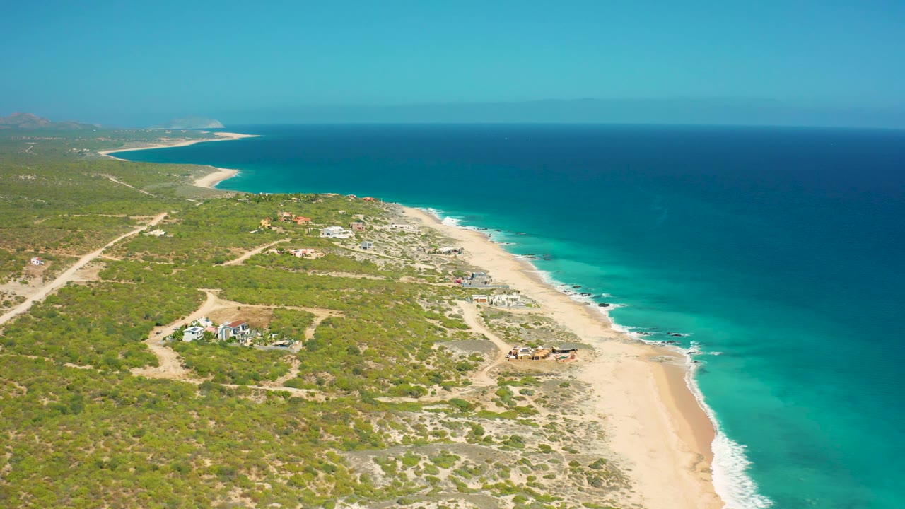 At 0:02, the El Cardoncito beach runs diagonally through the frame with turquoise water on the right, low green vegetation on the left, small inland structures, and no visible people.