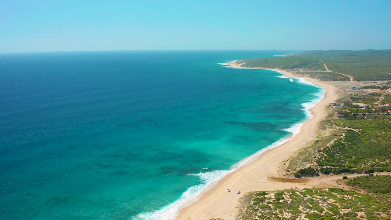 Aerial opening frame over El Cardoncito, Baja California Sur, with deep blue and turquoise sea on the left, a pale sandy beach curving along the coast, green scrubland to the right, and no visible people.