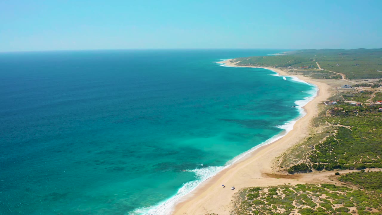 Aerial frame over El Cardoncito showing a broader band of turquoise shallows, white surf touching the sandy beach, sparse coastal buildings near the right edge, distant hills, and no visible people.
