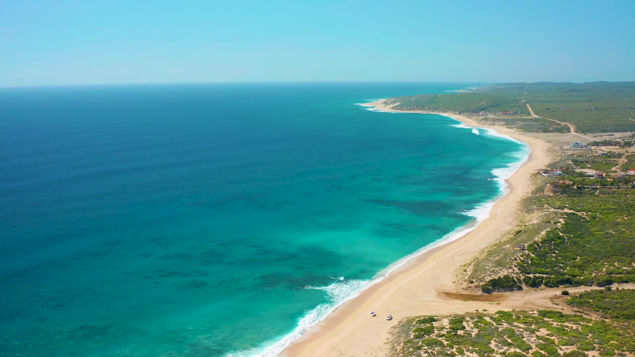 Aerial frame in El Cardoncito with the shoreline shifted slightly right, blue sea filling most of the left side, beach and surf running diagonally, small structures inland, and no visible people.