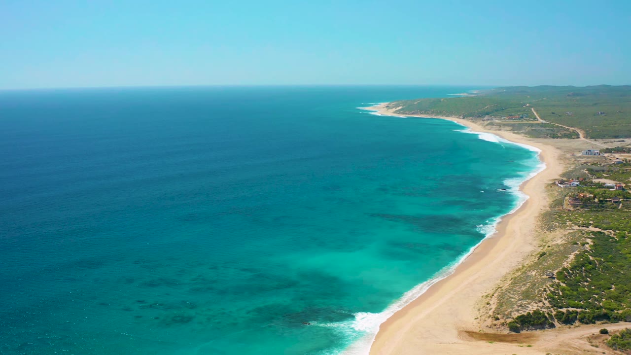 Wide aerial view in El Cardoncito showing deep blue offshore water, brighter turquoise shallows, a narrow white surf line, sandy beach, low green coastal land, and no visible people.