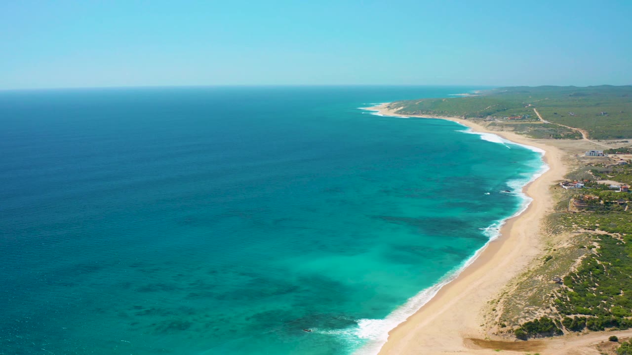 Drone-style view over the Baja California Sur coast with a long sand strip on the right, turquoise water across the center, darker reef-like patches under the water, green land in the background, and no visible people.