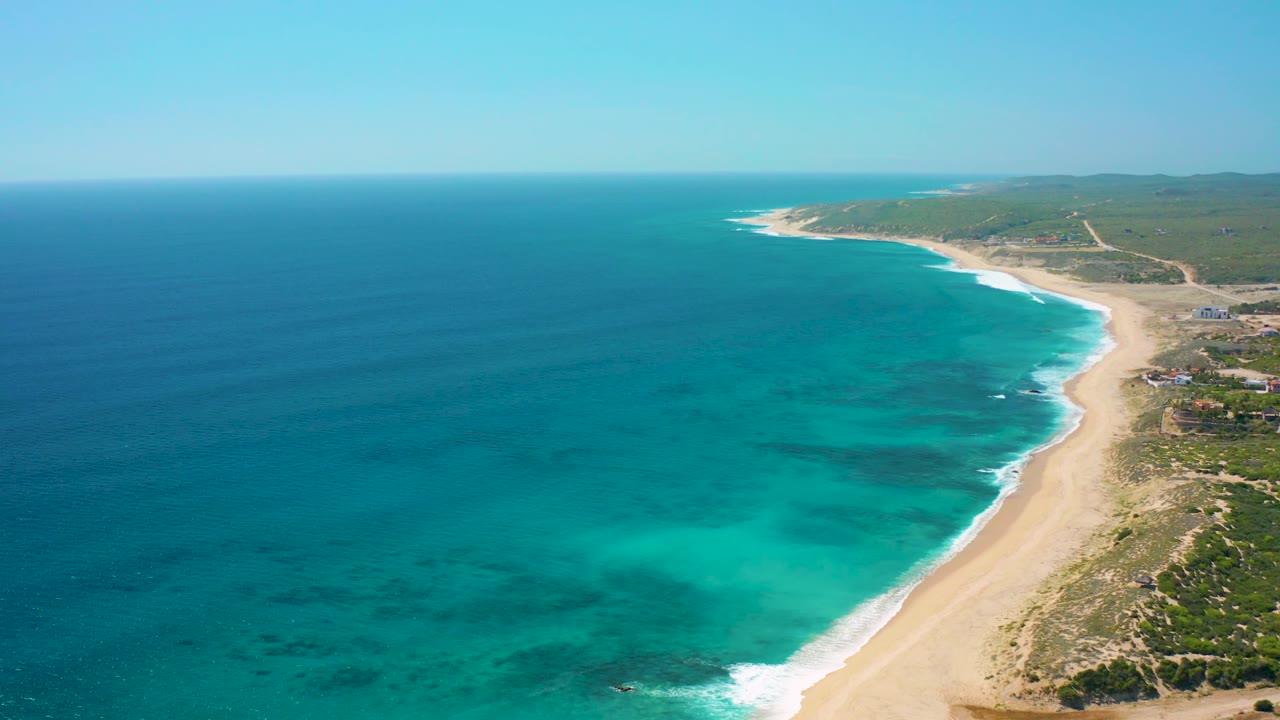 High aerial composition over El Cardoncito with open ocean dominating the left side, a curving sandy beach on the right, low vegetation inland, small distant structures near shore, and no visible people.