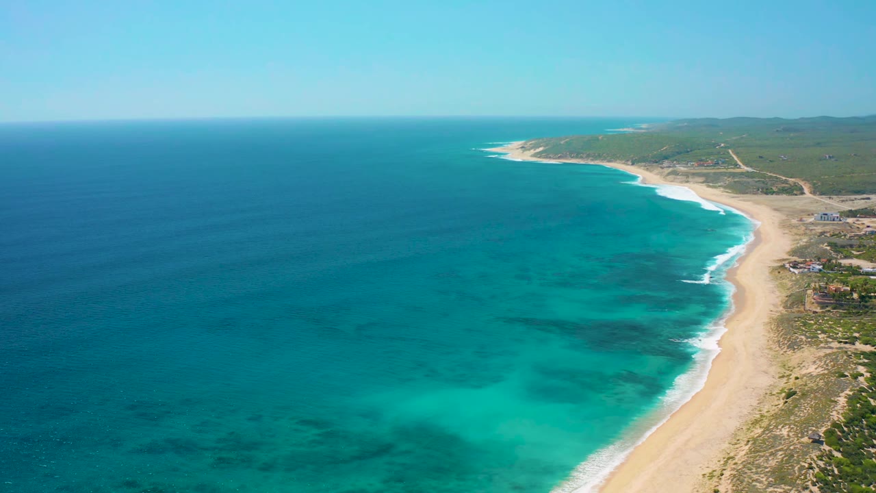 Drone view along the El Cardoncito shoreline, showing turquoise water bands, a bright surf edge, tan sand, sparse vegetation, distant tracks, and no visible people.