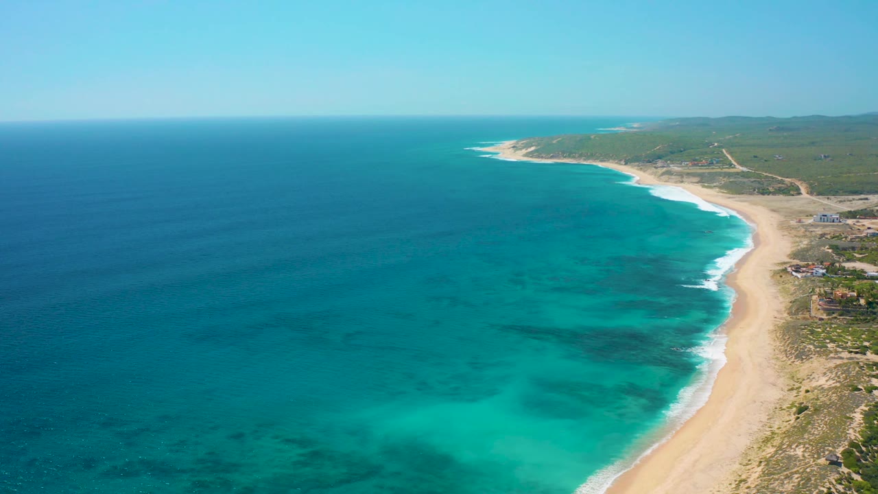 Aerial view of El Cardoncito coastline with clear blue-green water, darker reef or sea-bottom patches, white surf meeting the beach, open inland vegetation, and no visible people.