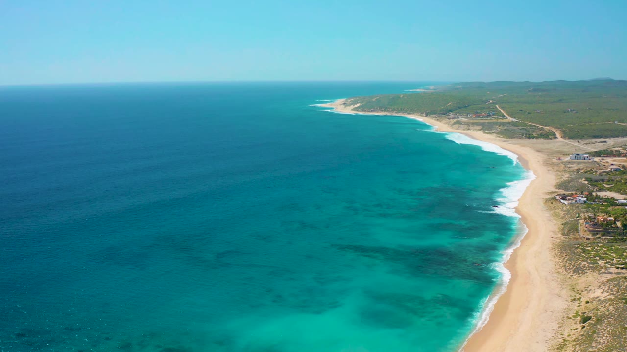 Drone-height view over El Cardoncito with deep blue water at left, brighter turquoise shallows near shore, a sandy beach on the right, and no visible people.