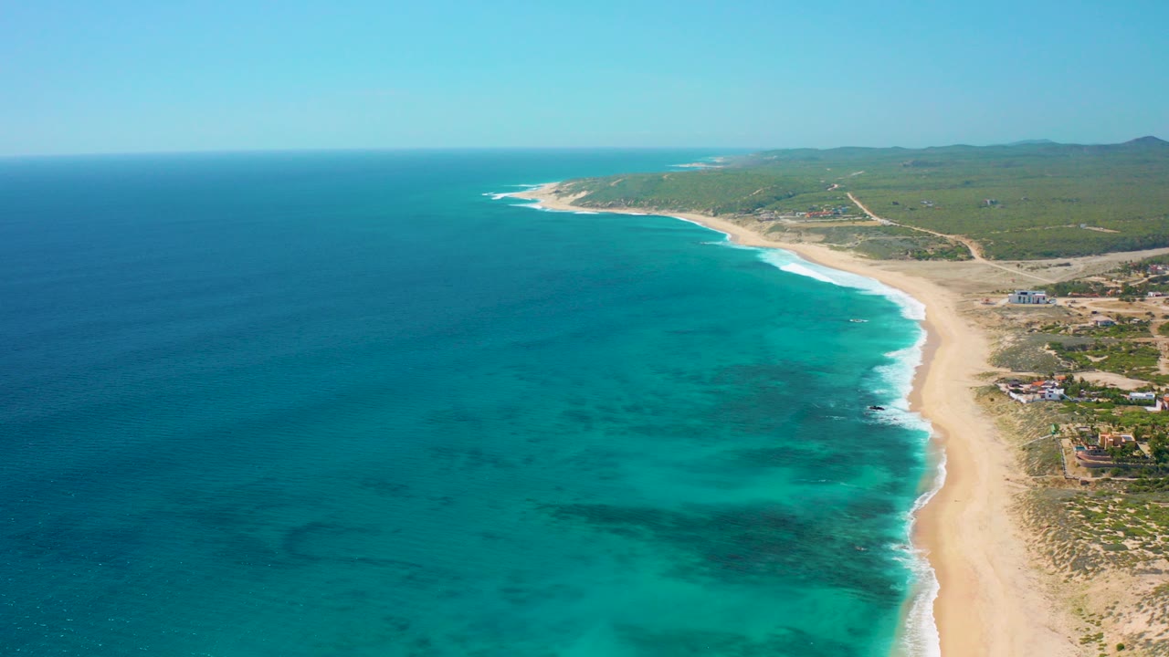 Aerial frame over El Cardoncito shows deep blue ocean shifting to turquoise shallows, a curved pale beach, inland scrub, scattered buildings, distant low hills, and no visible people.