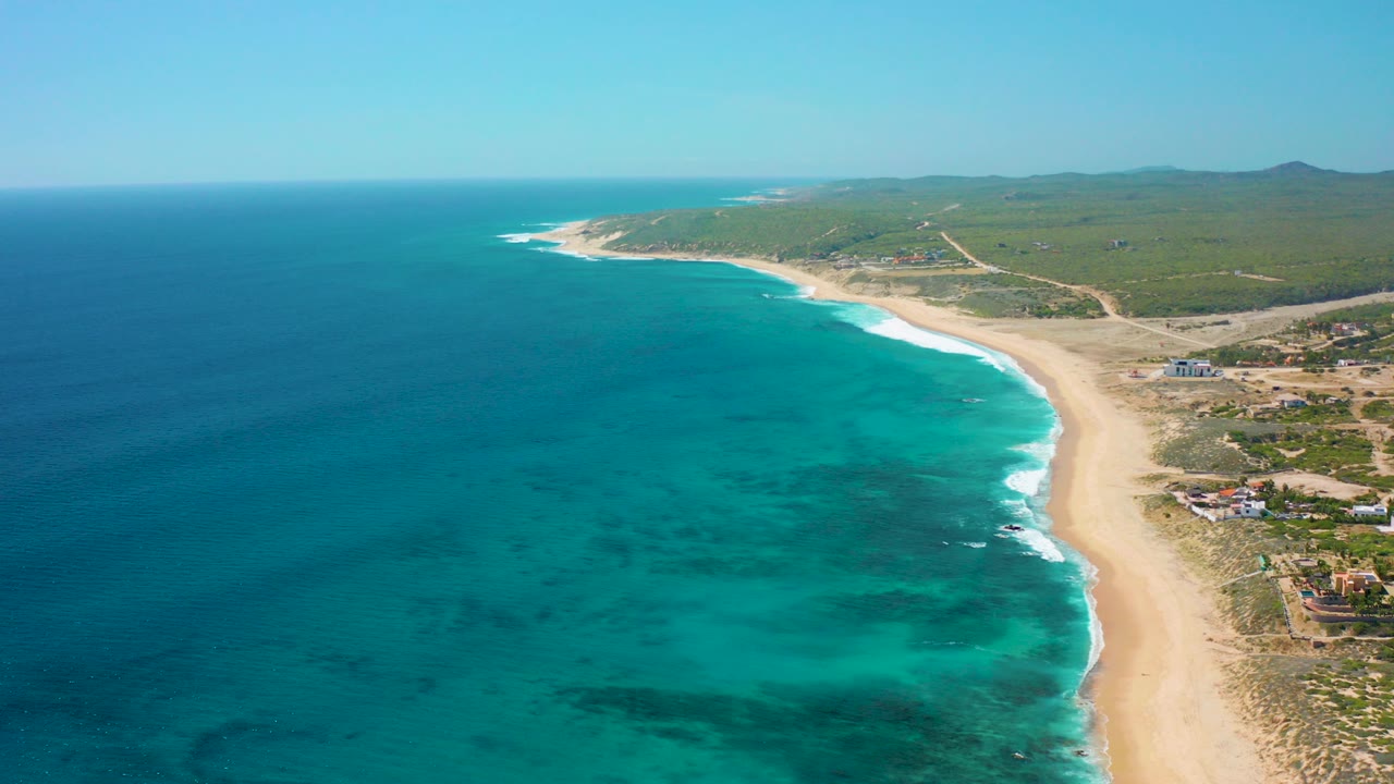 Opening aerial frame over El Cardoncito, Baja California Sur, Mexico, with deep blue ocean left, turquoise shallows near the beach, pale sand on the right, inland scrubland beyond, and no visible people.