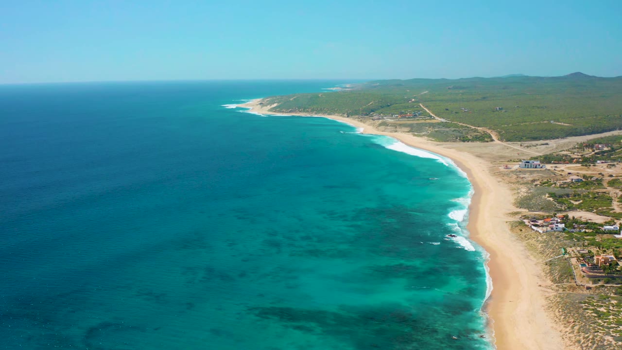 Aerial view in El Cardoncito with the ocean filling the left side, a bright surf edge tracing the beach, low inland vegetation and roads in the background, and no visible people.