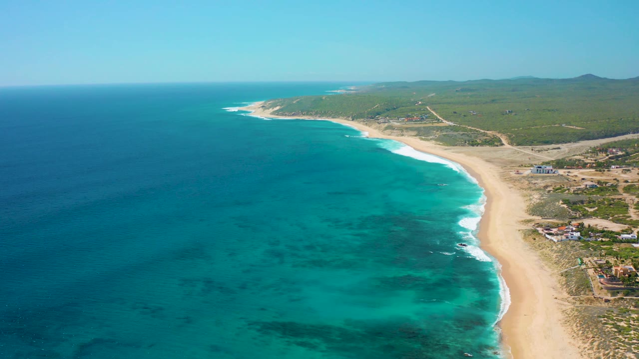 Aerial frame over El Cardoncito showing the beach curving along clear turquoise water, white waves on the shore, green terrain and roads inland, distant low hills, and no visible people.