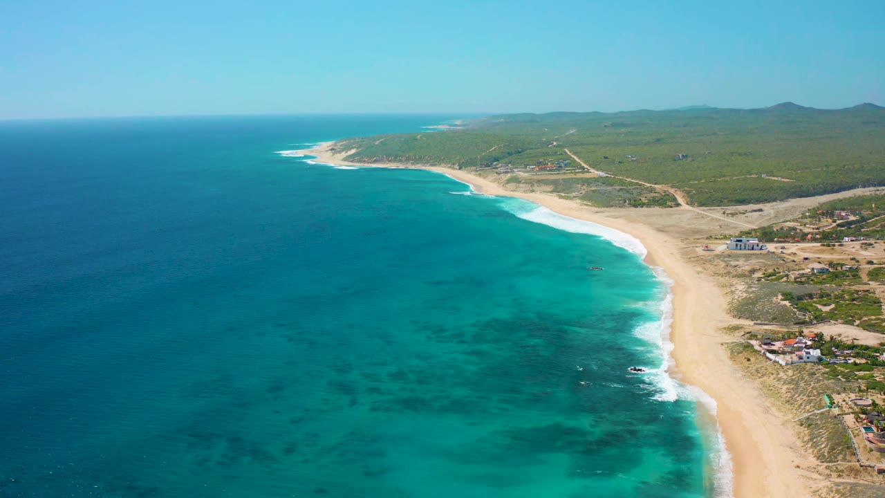 Aerial frame over El Cardoncito showing the shoreline bending into the distance, darker reef-like patches under the water, low green vegetation inland, and no visible people.