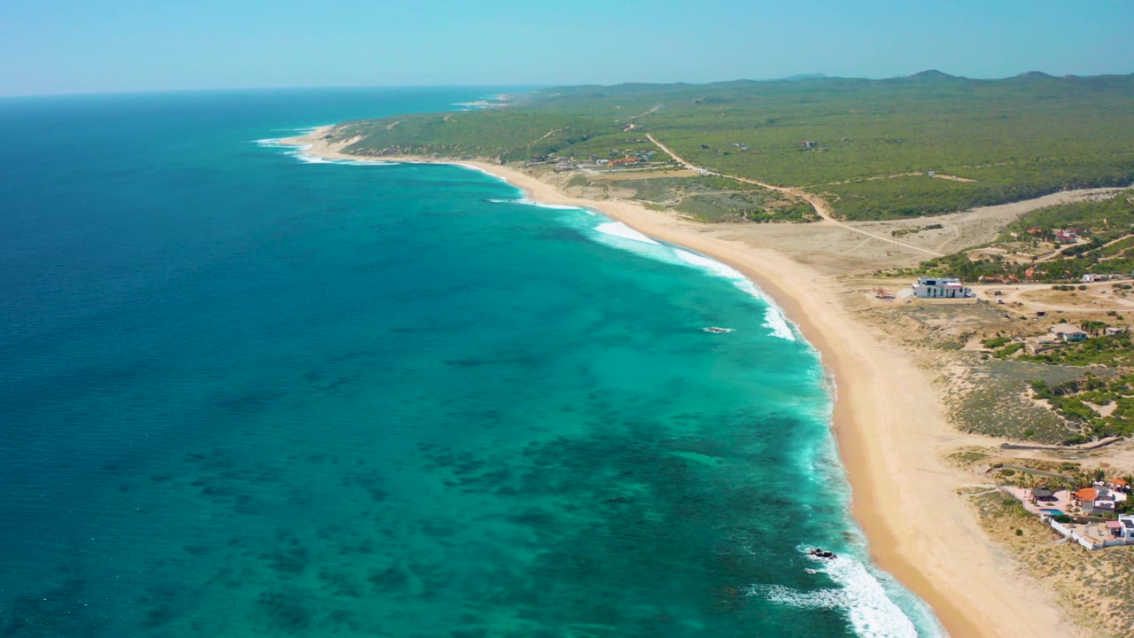 Aerial view over El Cardoncito, Baja California Sur, with turquoise ocean on the left, a wide sandy beach curving through frame, inland green brush, dirt pathways, scattered buildings, and no visible people.