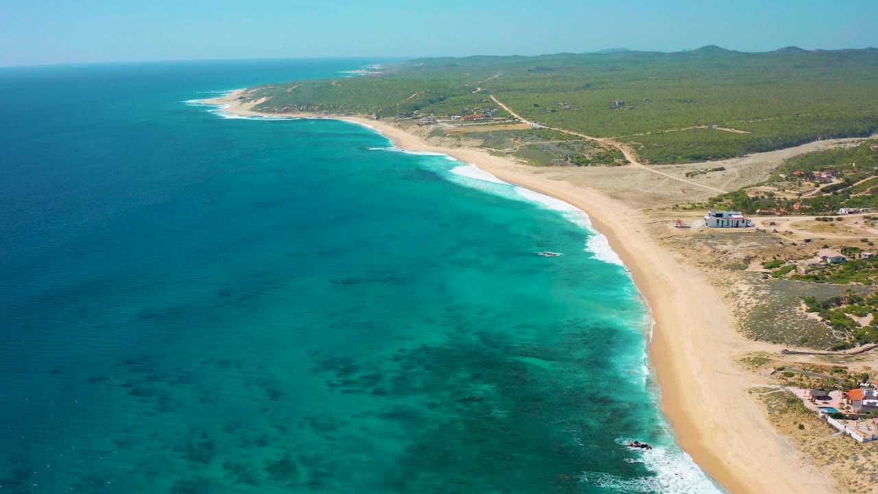 Wide aerial coastline with ocean at left, beach at center, and green land behind.