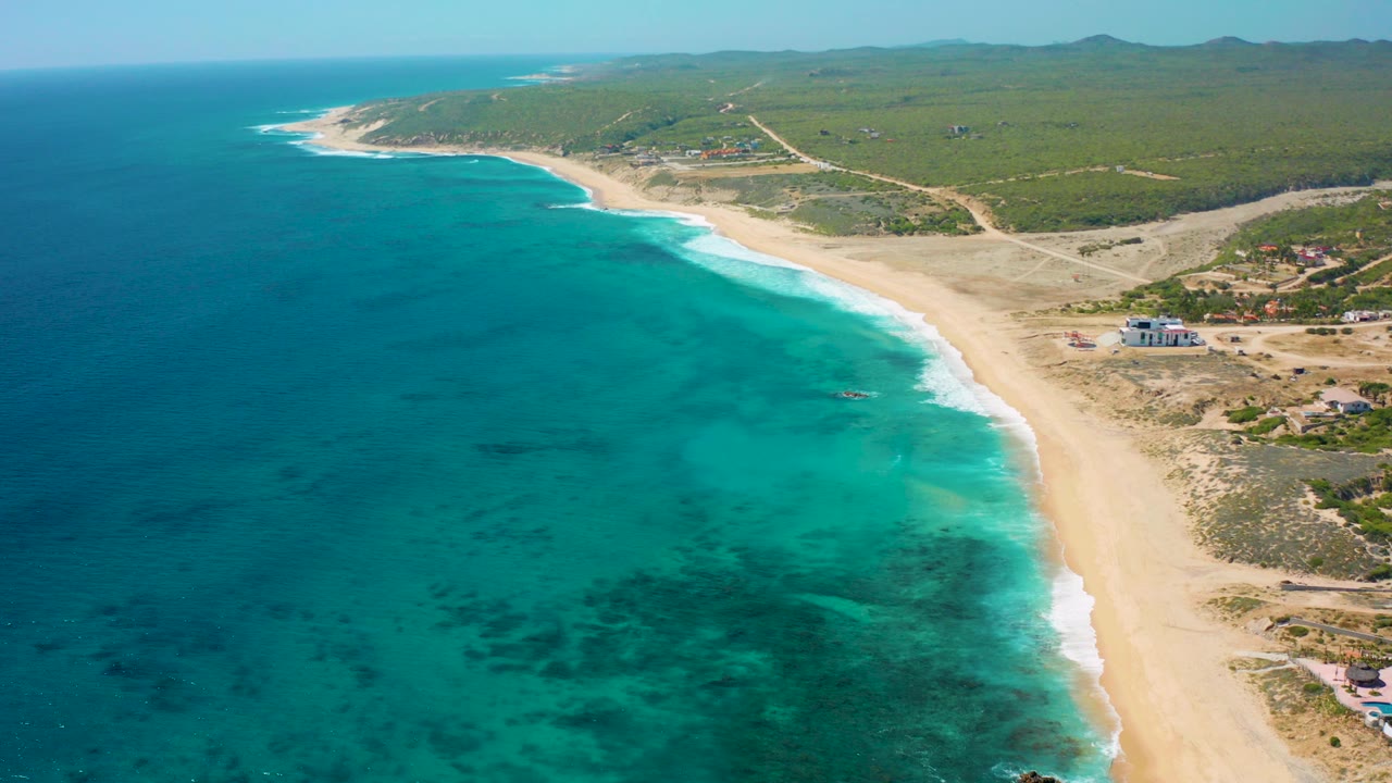 Opening aerial frame over El Cardoncito, Baja California Sur, showing turquoise water, pale sand, inland scrub, dirt roads, scattered buildings, and no visible people.