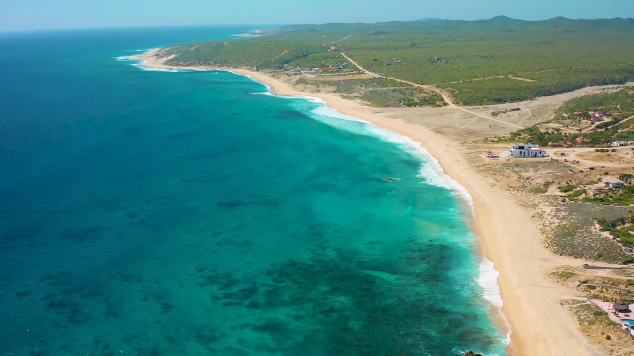 Aerial frame over El Cardoncito with the shoreline centered, darker patches under the blue sea, a wide sand strip, inland roads, sparse buildings, and no visible people.