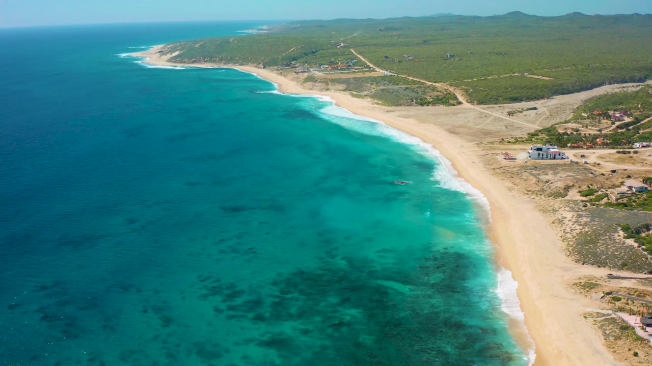 Aerial view in El Cardoncito, Baja California Sur, with the beach sweeping diagonally, clear blue shallows, coastal scrub inland, a few buildings, and no visible people.