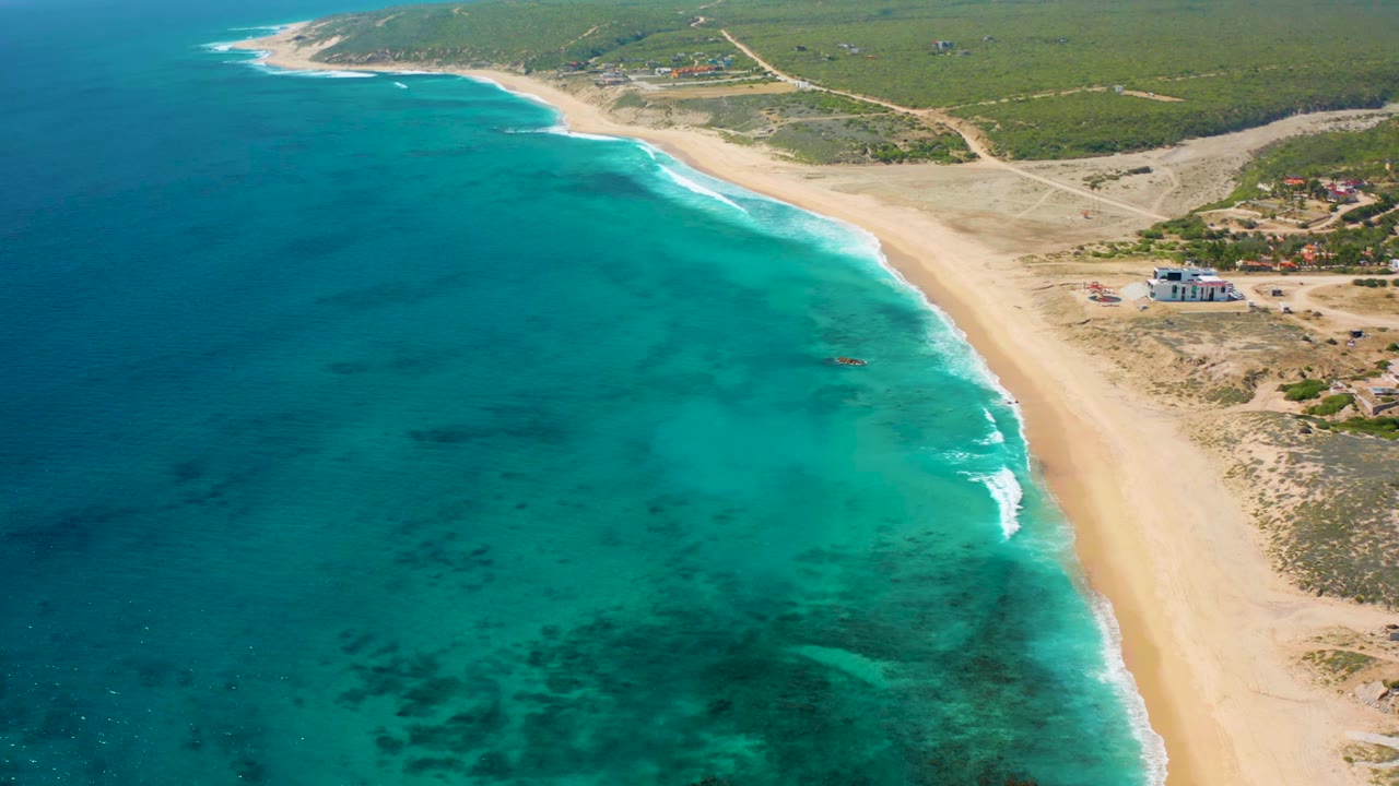 Elevated drone view of the El Cardoncito coast as blue-green water meets a wide sandy beach, with a small light-colored building and low vegetation inland, and no visible people.