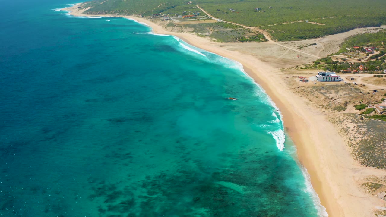 Aerial frame over El Cardoncito showing turquoise sea, darker underwater patches, a narrow band of white surf, pale sand, scrubland, dirt roads, and no visible people.
