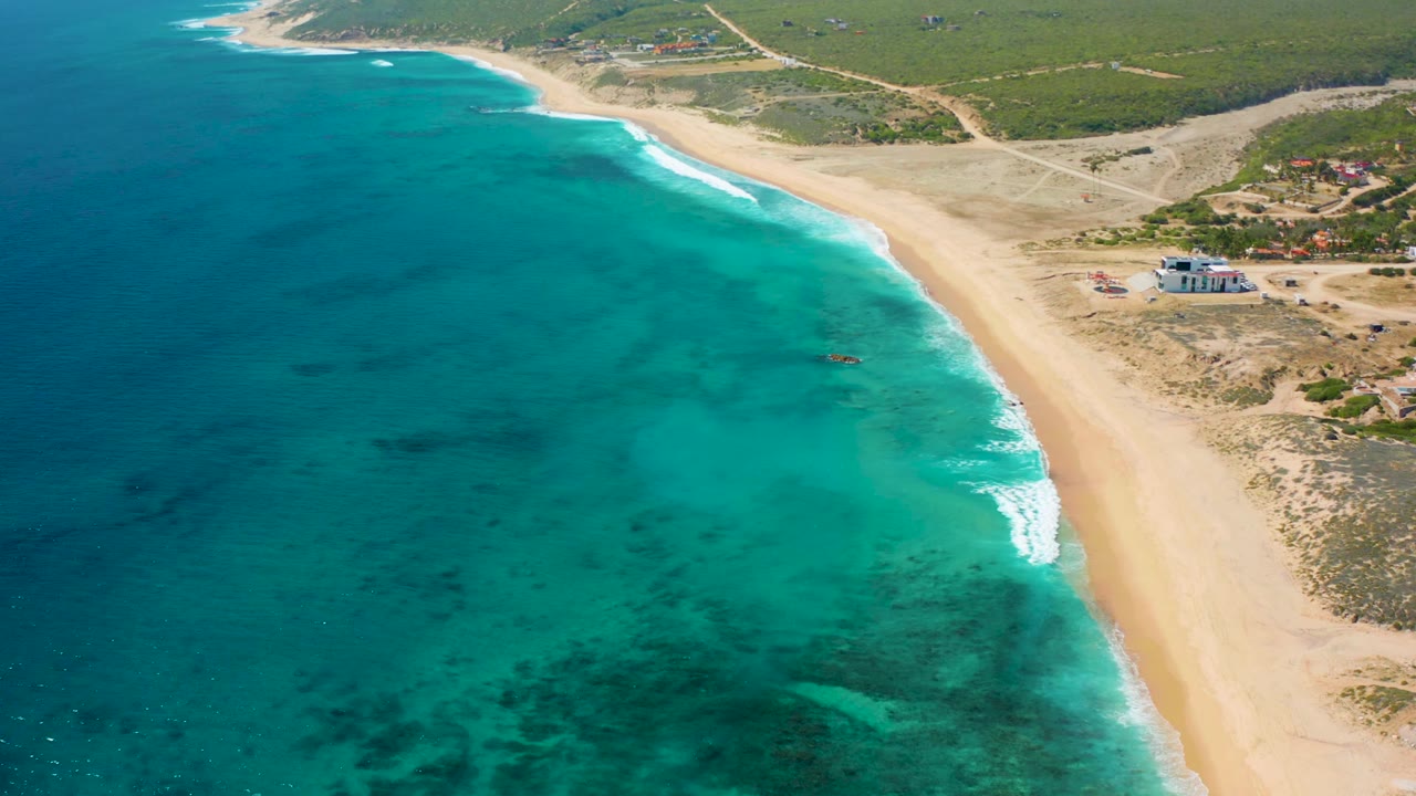 High oblique view at El Cardoncito with clear water across the left side, foamy waves along the sand, a light building near the beach, low vegetation inland, and no visible people.