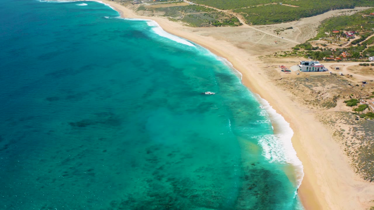 Aerial frame over El Cardoncito, Baja California Sur, showing turquoise shallows, a diagonal sand beach, inland scrub, a small offshore boat, and no visible people.