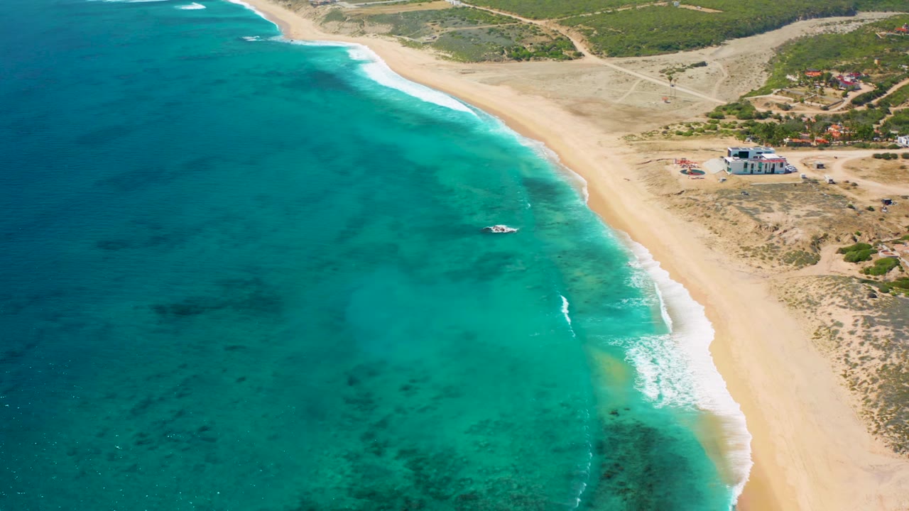 Aerial frame from El Cardoncito showing mild waves reaching the sand, turquoise water with darker submerged areas, inland scrubland, and no visible people.