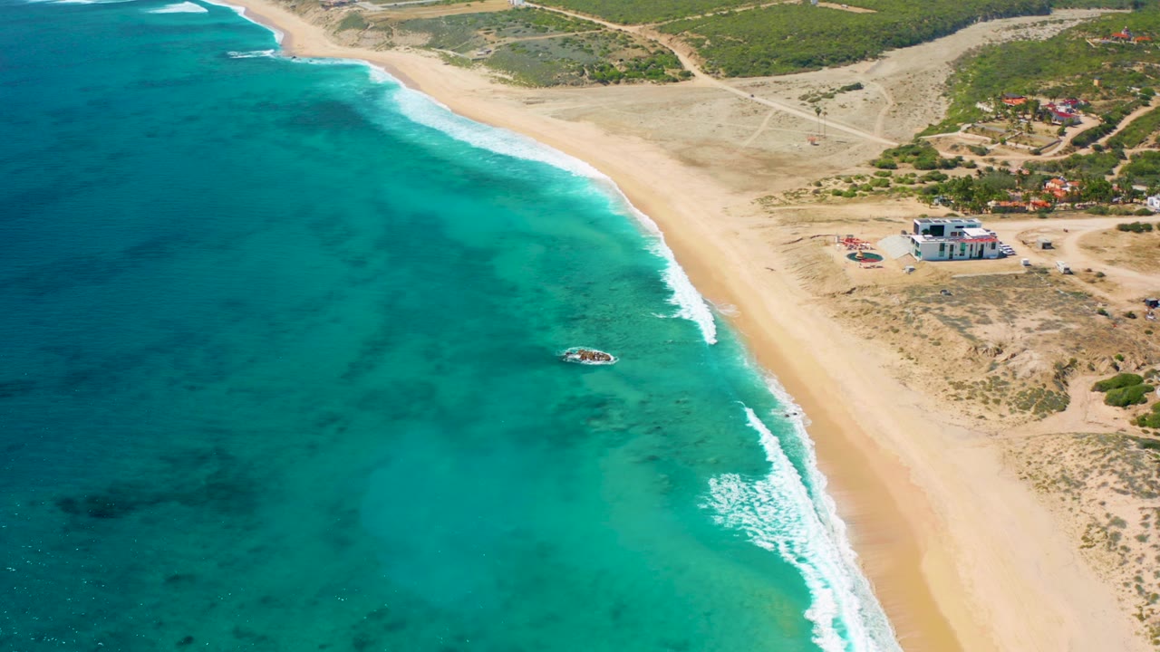 High aerial view over El Cardoncito, Baja California Sur, with turquoise water on the left, a pale sand beach curving through the frame, inland scrub and buildings on the right, and no visible people.