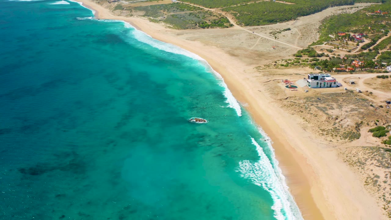 Drone view of clear water, surf, sand, and scattered shore buildings.