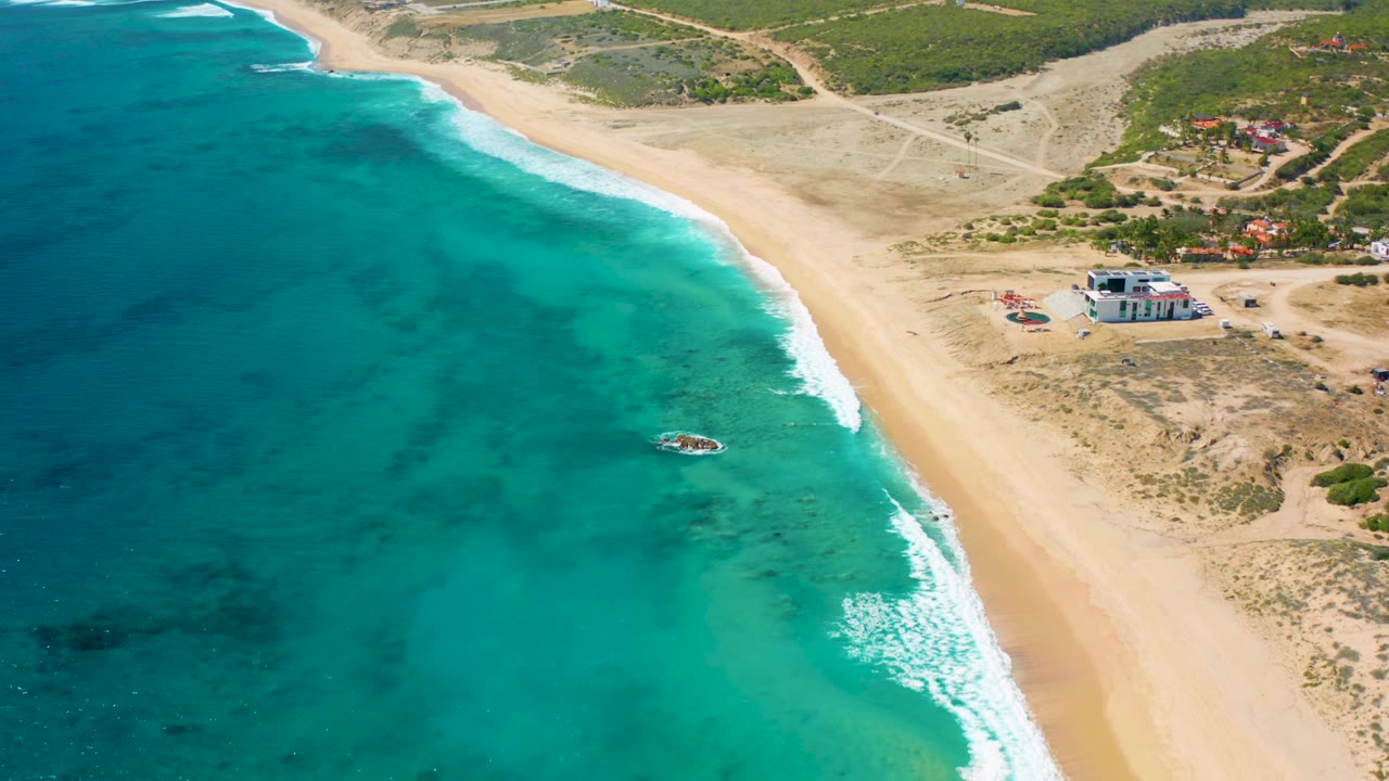 High-angle El Cardoncito coastline frame with darker underwater patches in turquoise sea, a white surf line on the sand, dry land and buildings beyond the beach, a small boat near the shallows, and no visible people.