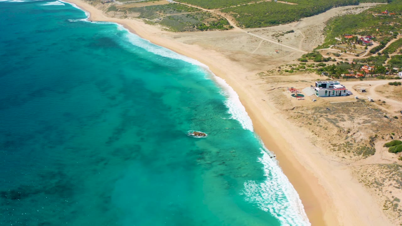 Wide aerial beach view with surf, turquoise water, and inland dirt tracks.