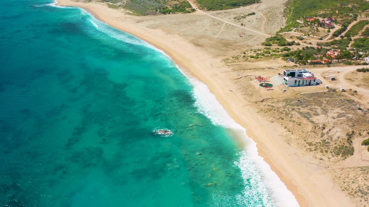 Aerial frame over El Cardoncito, Baja California Sur, showing turquoise water beside a pale beach, a small boat offshore, dry scrubland, low buildings, and no visible people.