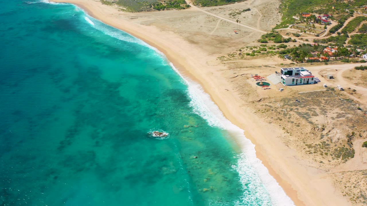 El Cardoncito aerial frame showing darker reef-like patches under the water, a small boat near the surf, pale sand, inland buildings, and no visible people.