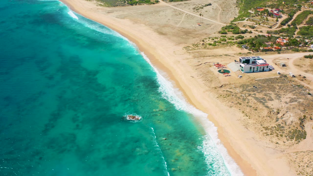 Overhead coastal view with beach, surf, boat, scrubland, and a low shelter building.