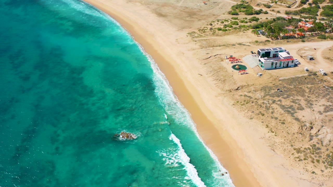 Aerial frame over El Cardoncito with turquoise water on the left, a pale sandy beach through the center, a white beachfront house near the dunes, and no visible people.