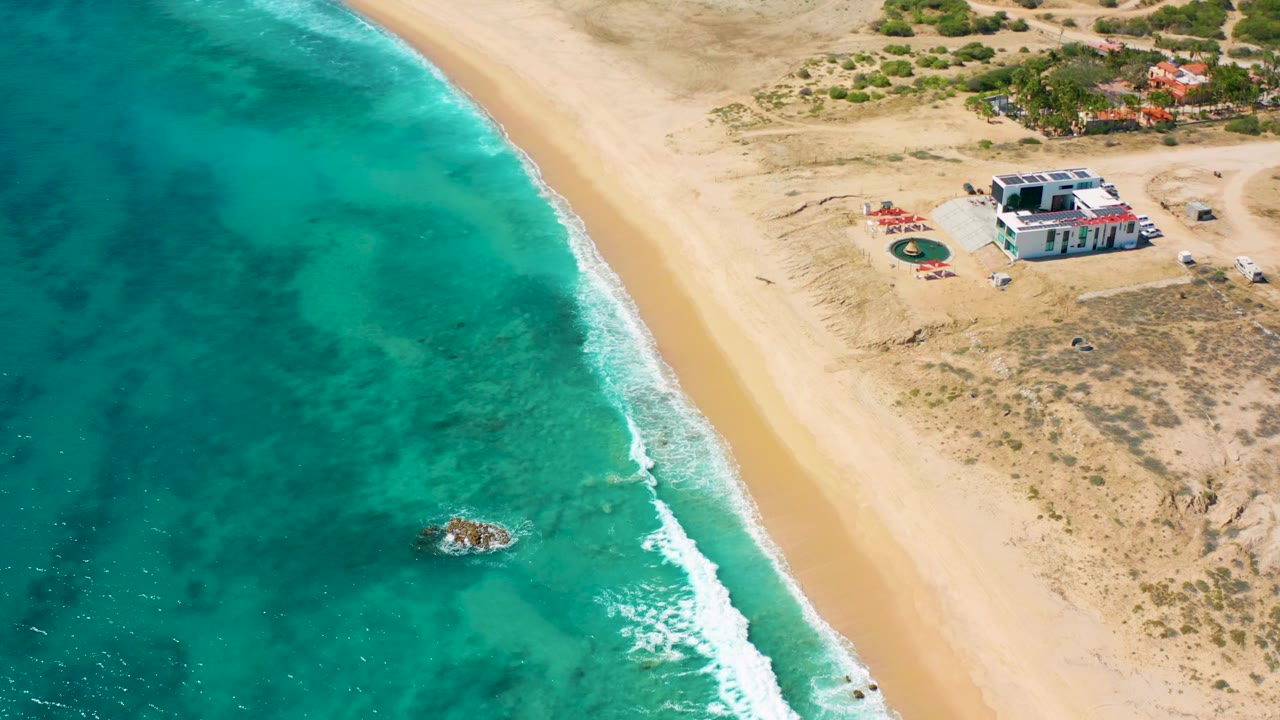 El Cardoncito aerial frame with darker patches in the turquoise water, white foam along the shore, open sand, a white house above the beach, and no visible people.