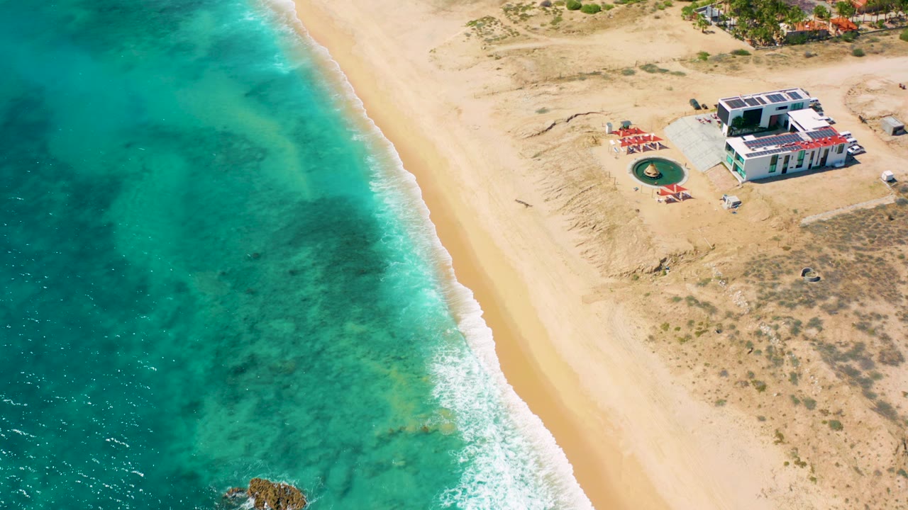 High aerial view over El Cardoncito, Baja California Sur, with turquoise ocean at left, pale beach at center, a small structure near the upper right, and no visible people.