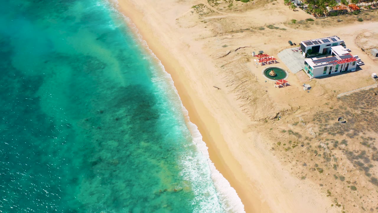 Drone-like view over El Cardoncito with blue-green water filling the left side, pale empty sand through the middle, beachside structures at upper right, and no visible people.