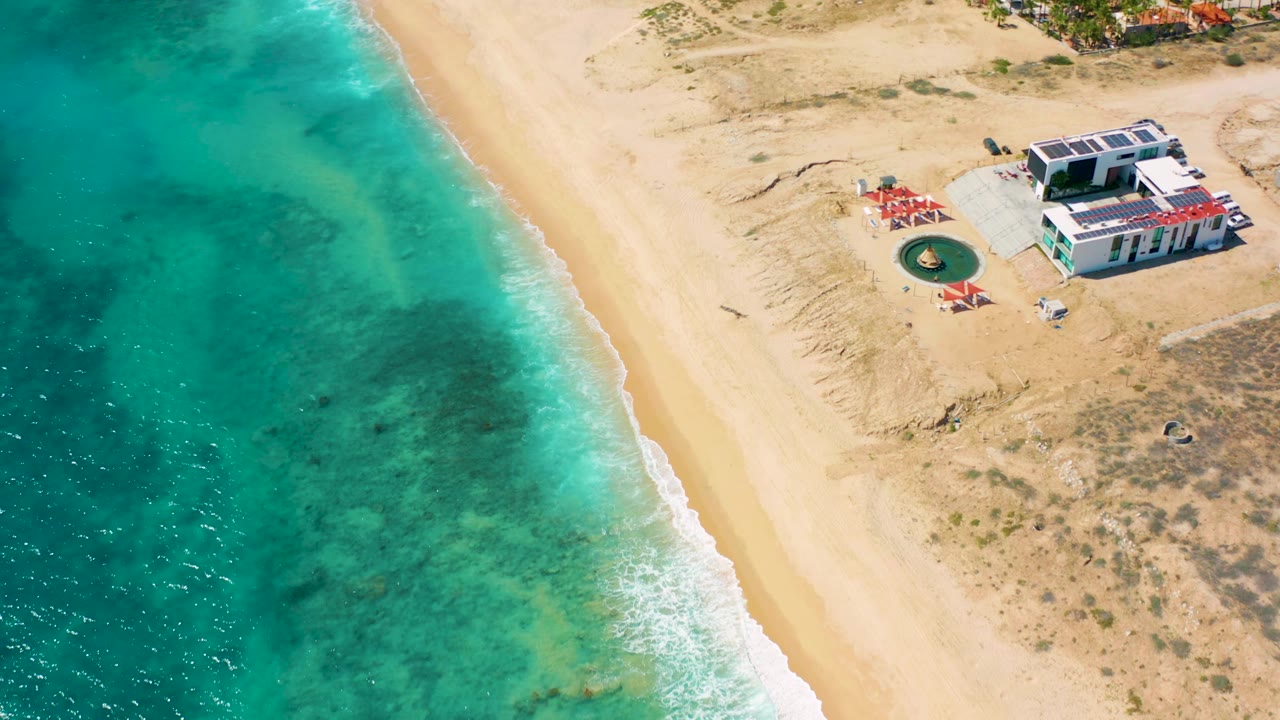 Top-down beach view with turquoise water, surf, dry vegetation, and a small structure.
