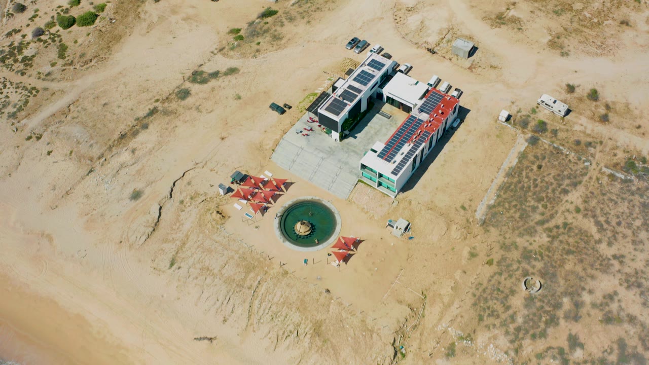Opening aerial frame over El Cardoncito, Baja California Sur, with a modern beachside building, red shade structures, a round green water feature, dry scrubland, pale sand, and no visible people.