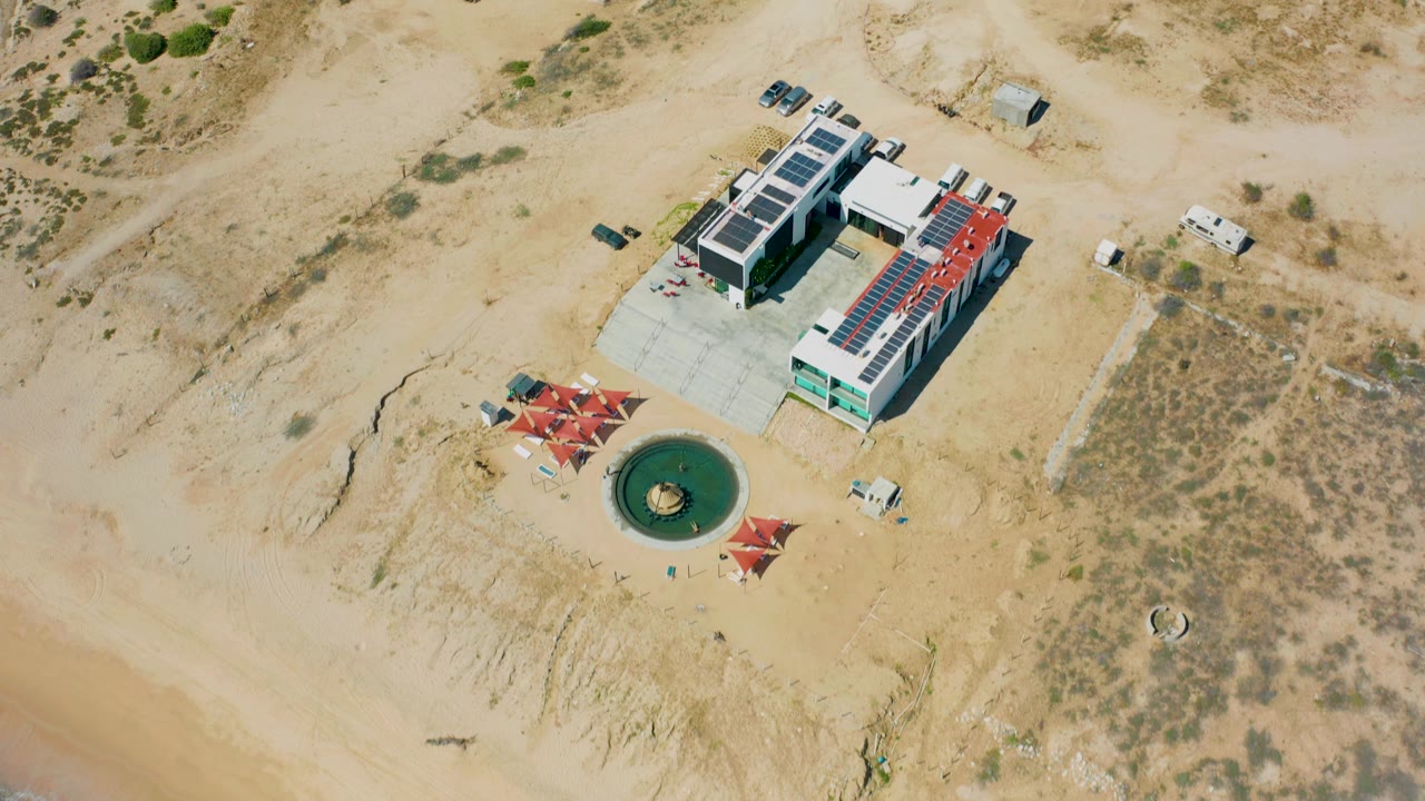 Aerial frame in El Cardoncito showing the beachside structure from above, with dirt access roads behind it, dry brush to the right, red shade canopies near a circular green feature, and no visible people.