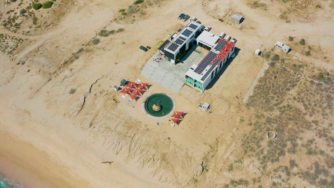 The third frame holds the El Cardoncito building near the top of the view, with a circular green water feature and red shade structures below it, sandy terrain around it, and no visible people.