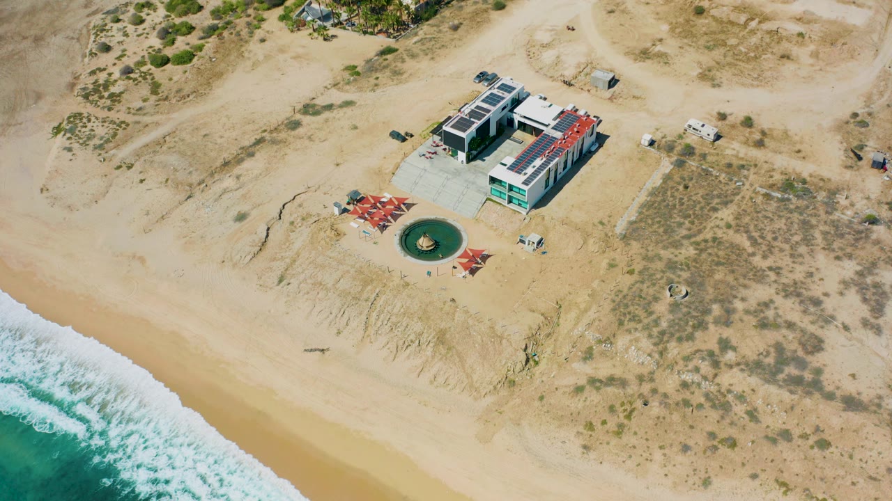 Aerial frame of a modern beachfront house in El Cardoncito, Baja California Sur, with turquoise surf at lower left, dry scrub inland, and no visible people.