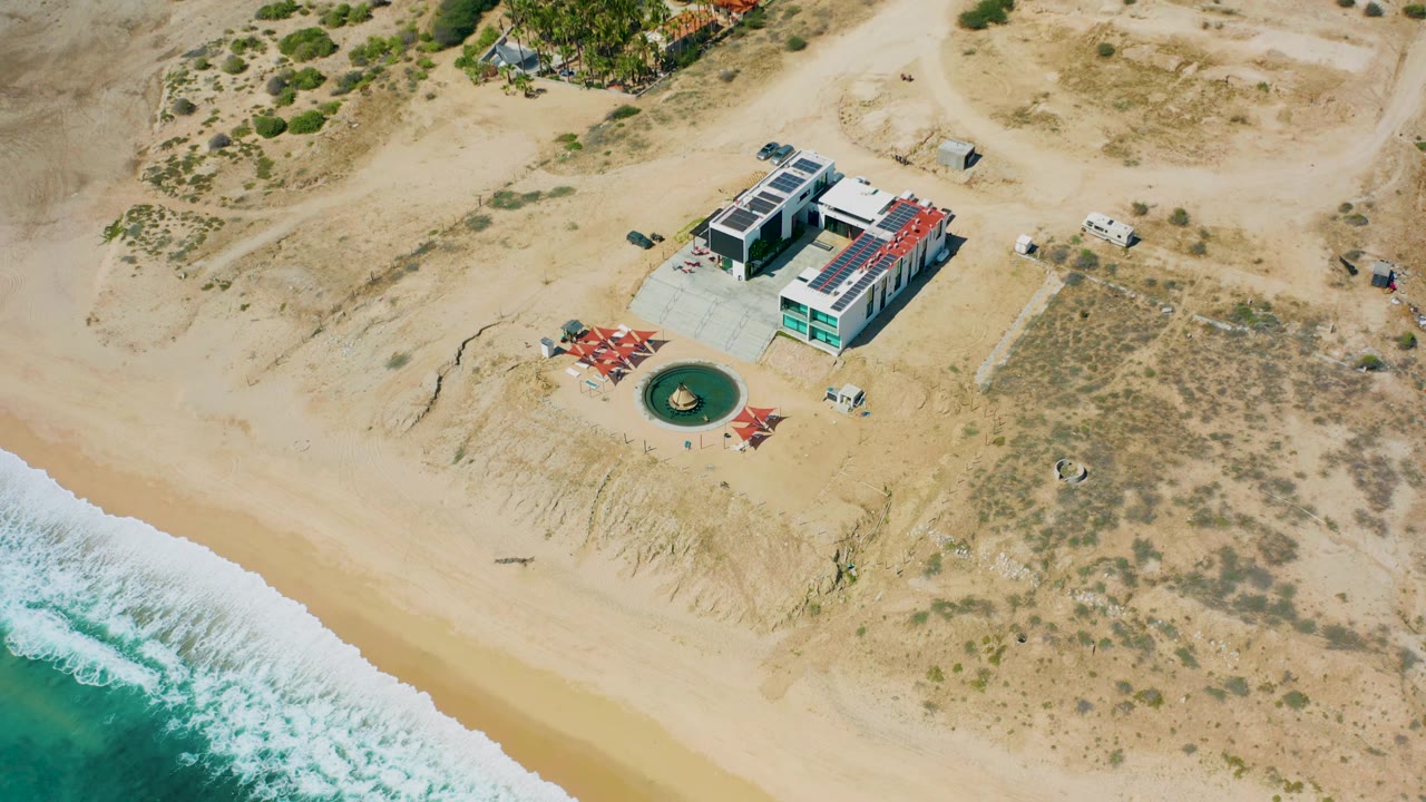 Aerial view of the El Cardoncito beach house with pale sand in front, a band of white surf, sandy tracks inland, and no visible people.