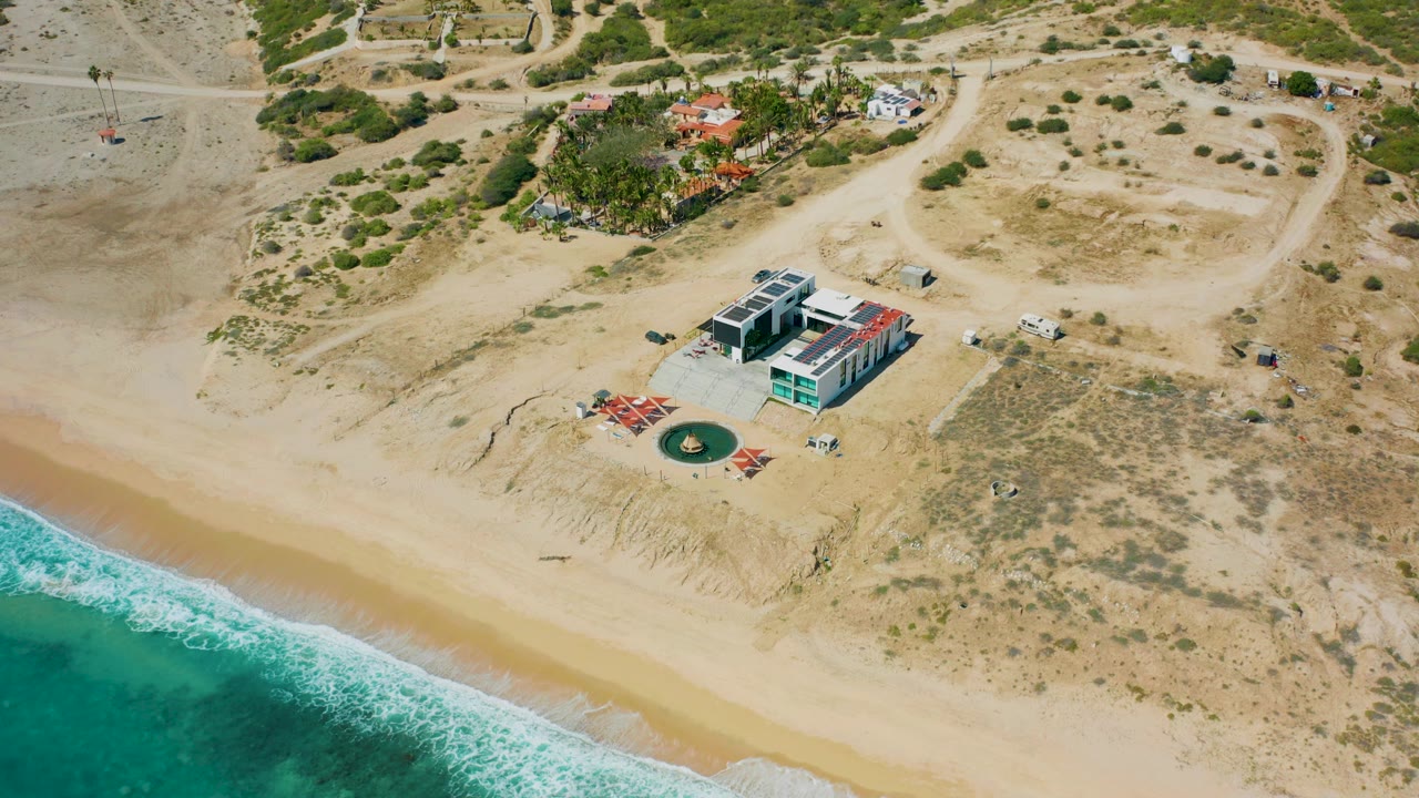 High aerial frame over El Cardoncito showing a modern beachfront house, pale sand, turquoise water at the lower left, dry scrubland, dirt roads, and no visible people.