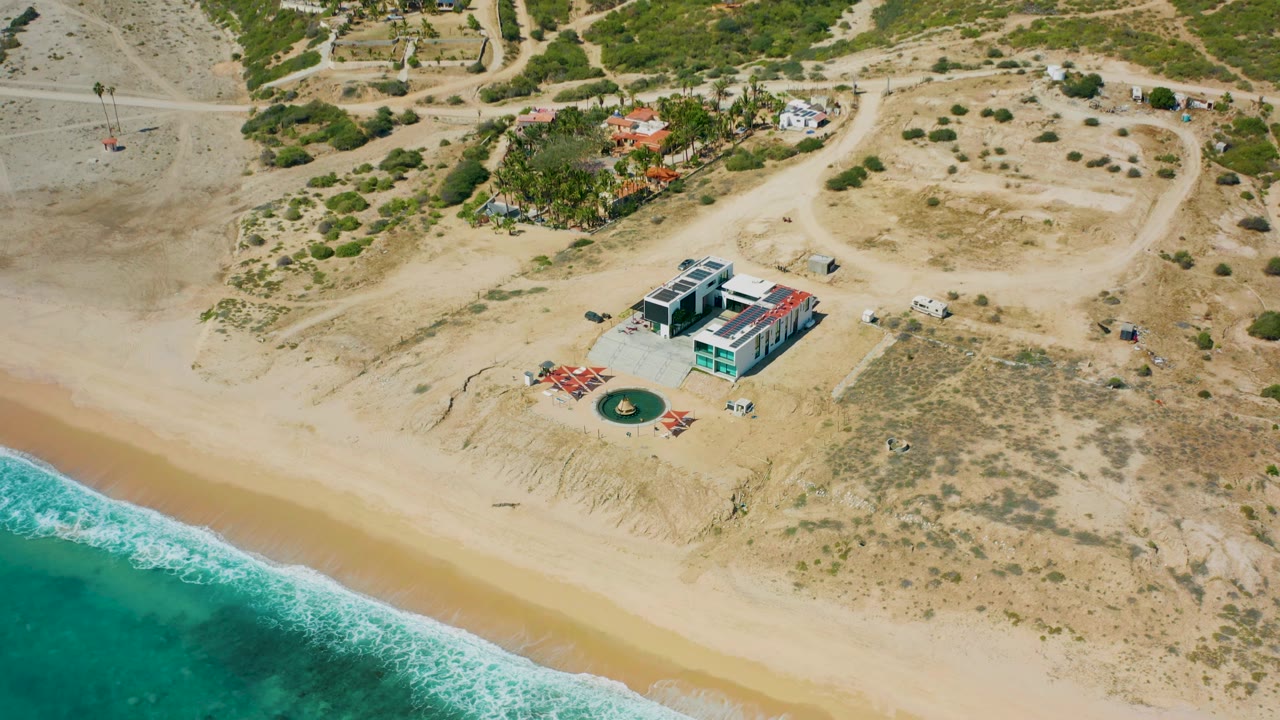High-angle aerial frame over El Cardoncito with a modern mansion near the shoreline, bright turquoise water, tan beach, scrub-covered ground, dirt access roads, and no visible people.